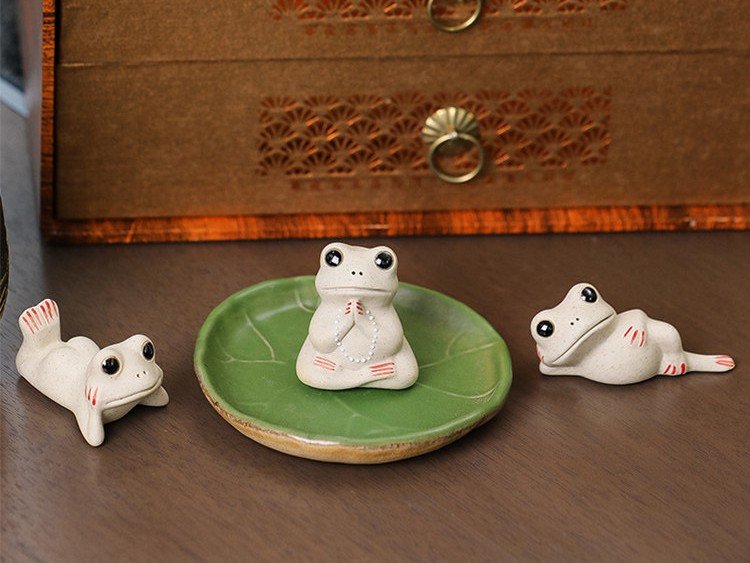 Three ceramic frog tea pets in different poses with green leaf tray on wooden table