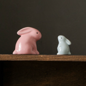 Two minimalist ceramic rabbit tea pets in pink and blue displayed on wooden shelf