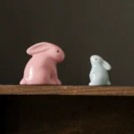 Two minimalist ceramic rabbit tea pets in pink and blue on wooden shelf